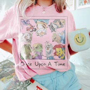 May include: Pink t-shirt featuring a square graphic with cartoon characters and the text "Once Upon A Time." A white mug with a yellow smiley face is held in the hand. Light blue shorts are also visible.