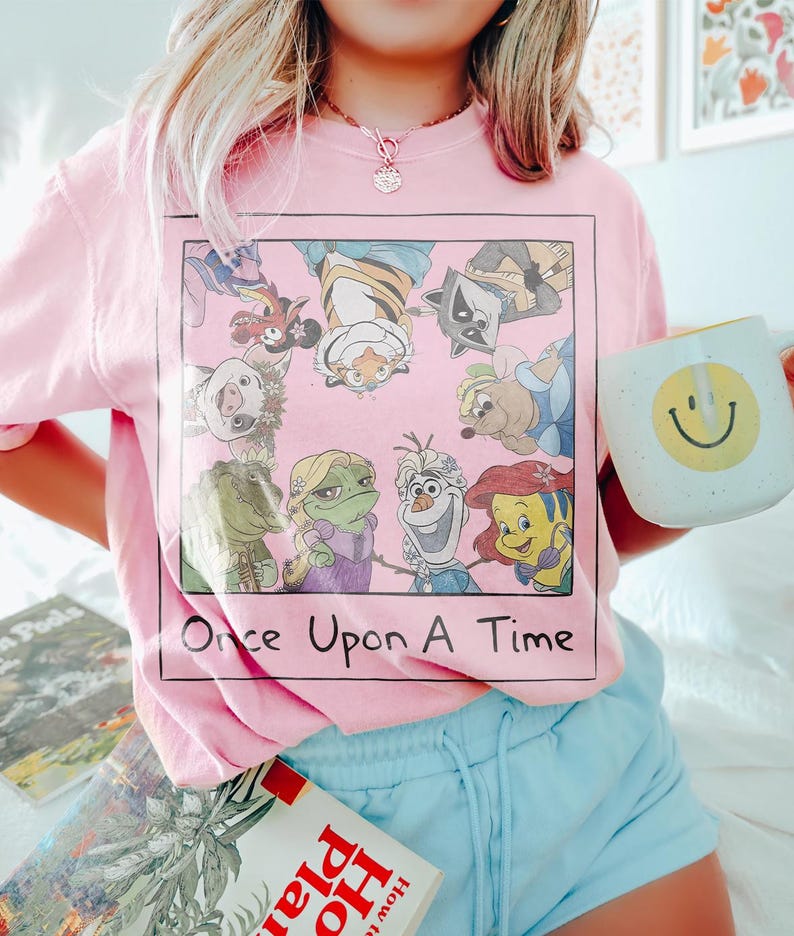 May include: Pink t-shirt featuring a square graphic with cartoon characters and the text "Once Upon A Time." A white mug with a yellow smiley face is held in the hand. Light blue shorts are also visible.