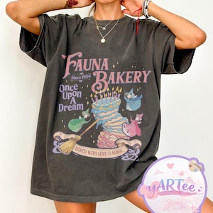 May include: Dark gray t-shirt featuring the text "Fauna Bakery Since 1959 Once Upon A Dream" with a whimsical illustration of a layered cake, fairies, and the phrase "Baked with Love & Magic".