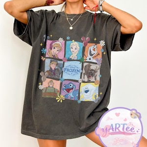 May include: Dark gray t-shirt featuring a colorful graphic of characters from the Disney movie "Frozen." The design includes images of Anna, Elsa, Olaf, Kristoff, Sven, and other characters, with the word "Frozen" in the center.