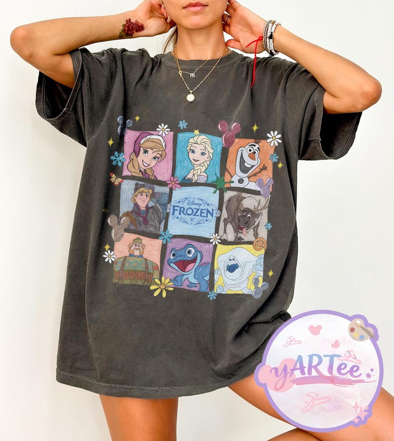 May include: Dark gray t-shirt featuring a colorful graphic of characters from the Disney movie "Frozen." The design includes images of Anna, Elsa, Olaf, Kristoff, Sven, and other characters, with the word "Frozen" in the center.
