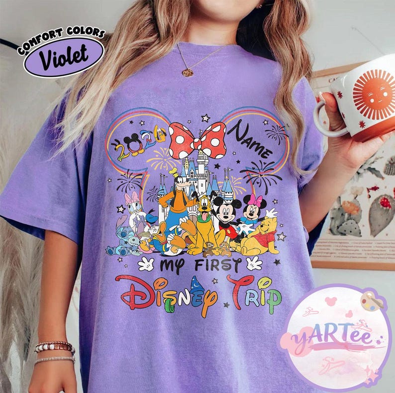 May include: Violet Comfort Colors t-shirt featuring a colorful graphic of Disney characters, fireworks, and a castle. The design includes the text "My First Disney Trip" and a customizable name area. The shirt has short sleeves.