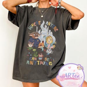 Puede incluir: Camiseta gris oscuro con un gr&aacute;fico colorido de personajes de animales animados y el texto "WHERE ANYONE CAN BE ANYTHING". El dise&ntilde;o incluye un oficial de polic&iacute;a, un zorro, un guepardo y un conejo. La camiseta tiene mangas cortas.