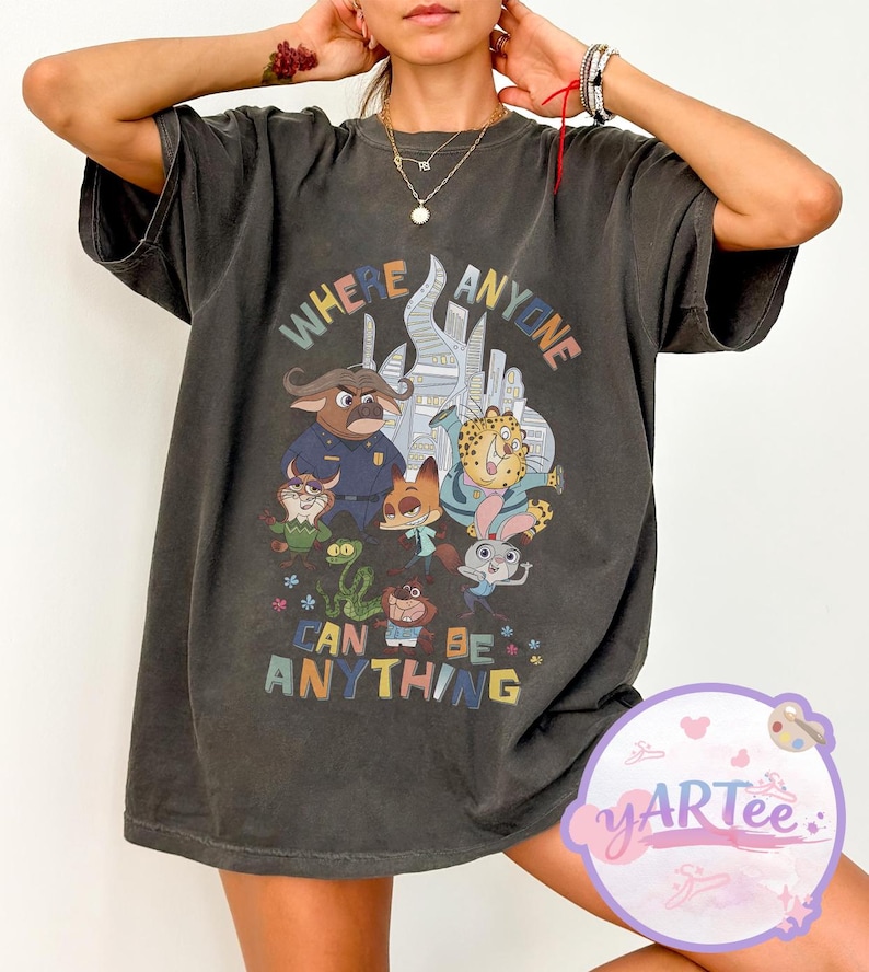 Puede incluir: Camiseta gris oscuro con un gr&aacute;fico colorido de personajes de animales animados y el texto "WHERE ANYONE CAN BE ANYTHING". El dise&ntilde;o incluye un oficial de polic&iacute;a, un zorro, un guepardo y un conejo. La camiseta tiene mangas cortas.