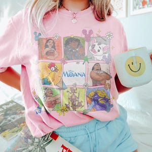 May include: Pink crew neck t-shirt featuring a colorful graphic of characters from the Disney movie Moana. The shirt includes the movie title and is paired with light blue shorts. A white mug with a yellow smiley face is held in the hand.