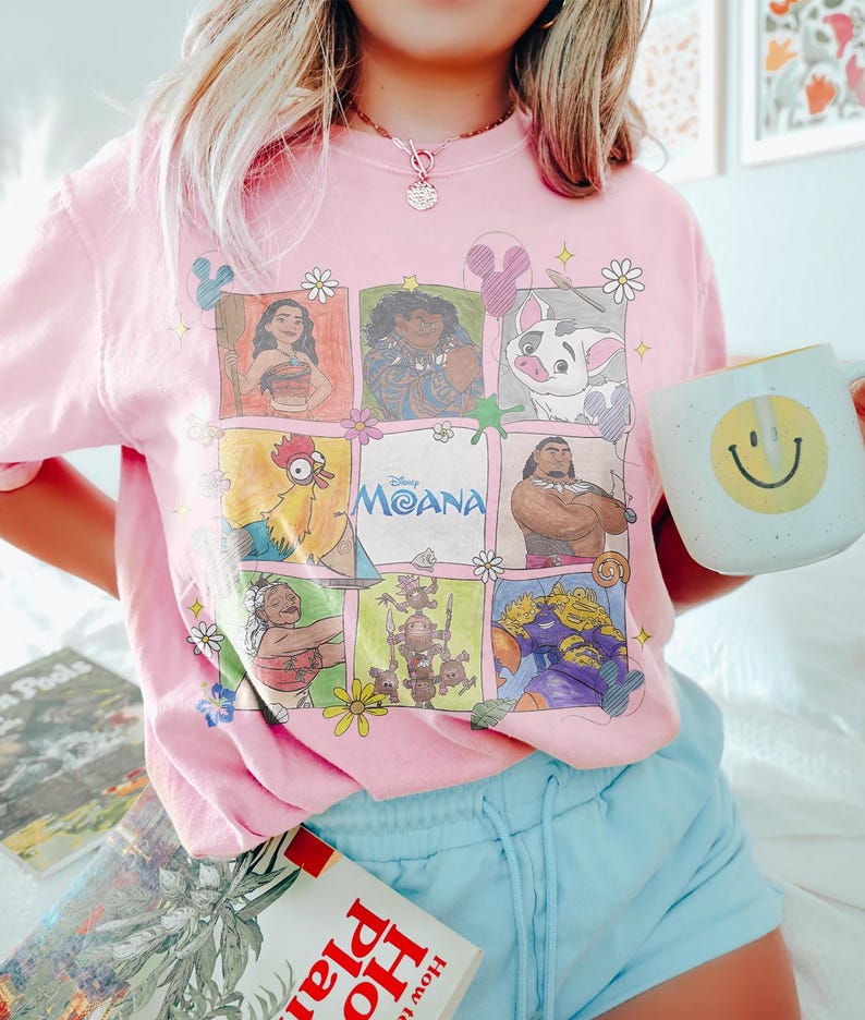 May include: Pink crew neck t-shirt featuring a colorful graphic of characters from the Disney movie Moana. The shirt includes the movie title and is paired with light blue shorts. A white mug with a yellow smiley face is held in the hand.