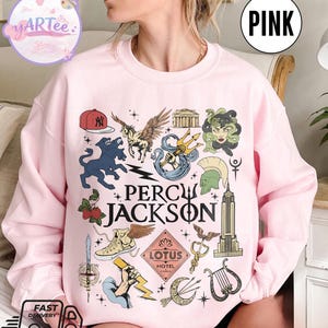 Pu&ograve; includere: Felpa rosa chiaro con un design grafico con la scritta "PERCY JACKSON" e varie illustrazioni. Il design include un berretto da baseball, un cavallo alato, un tridente e altri elementi mitologici.