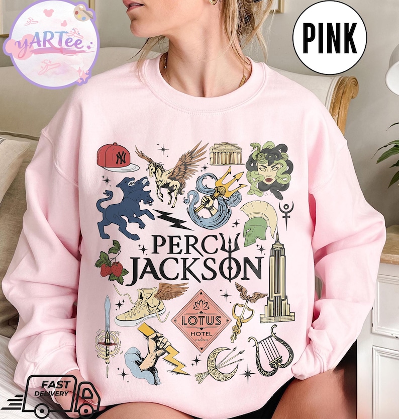 Pu&ograve; includere: Felpa rosa chiaro con un design grafico con la scritta "PERCY JACKSON" e varie illustrazioni. Il design include un berretto da baseball, un cavallo alato, un tridente e altri elementi mitologici.