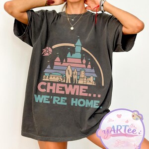 Puede incluir: Camiseta gris oscuro con un gráfico colorido que dice "CHEWE... WE'RE HOME." El diseño incluye un castillo, personajes y una nave espacial. La camiseta tiene mangas cortas y un corte holgado.