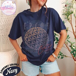 May include: Navy blue t-shirt with a graphic of the EPCOT Center ball, the word "EPCOT" above it, and "Winter 1982" below. The shirt is labeled "Comfort Colors Navy".
