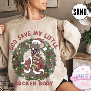 May include: Sand-colored sweatshirt with a graphic of a rat wearing a Santa hat and a scarf, holding a gift box. The text "GOD SAVE MY LITTLE BROKEN BODY" is printed above and below the graphic, surrounded by a Christmas wreath.