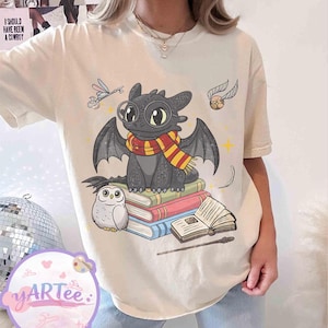 May include: Cream-colored t-shirt featuring a cartoon dragon wearing glasses and a scarf, sitting on books. A small owl, a key, a golden snitch, and a wand are also depicted. The text "I SHOULD HAVE BEEN A COWBOY" is visible.