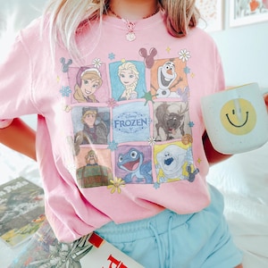 May include: A light pink t-shirt featuring a colorful graphic of characters from the Disney movie "Frozen." The design includes images of Anna, Elsa, Olaf, Kristoff, Sven, and other characters. A white mug with a yellow smiley face is held in the hand.
