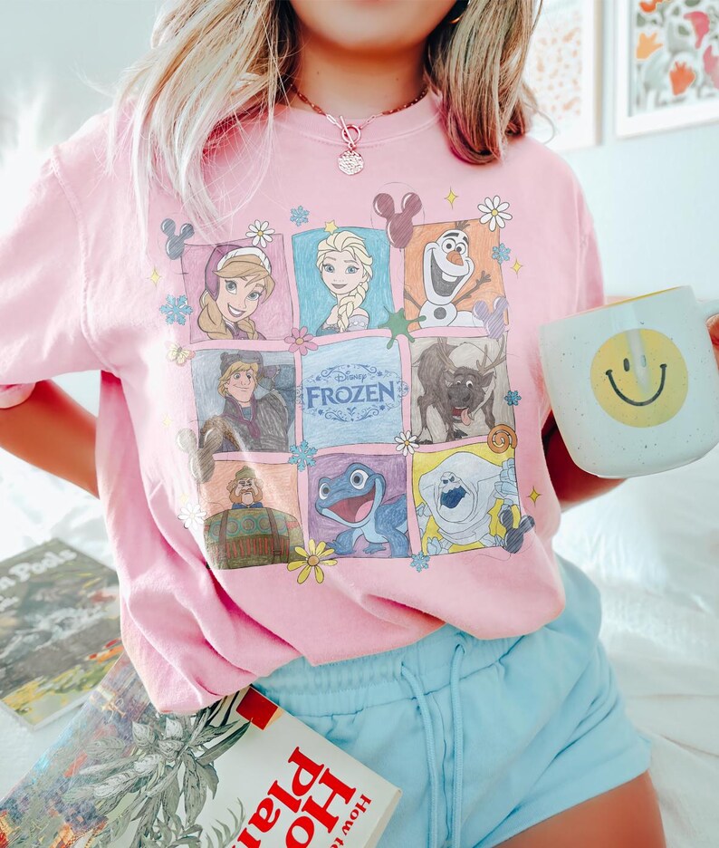 May include: A light pink t-shirt featuring a colorful graphic of characters from the Disney movie "Frozen." The design includes images of Anna, Elsa, Olaf, Kristoff, Sven, and other characters. A white mug with a yellow smiley face is held in the hand.
