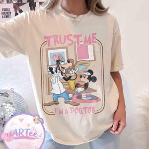 May include: Cream-coloured t-shirt with a cartoon illustration of Goofy and Mickey Mouse as doctors. The text "TRUST-ME I'M A DOGTOR" is printed in pink and brown. The shirt has a relaxed fit and is made of a soft material.