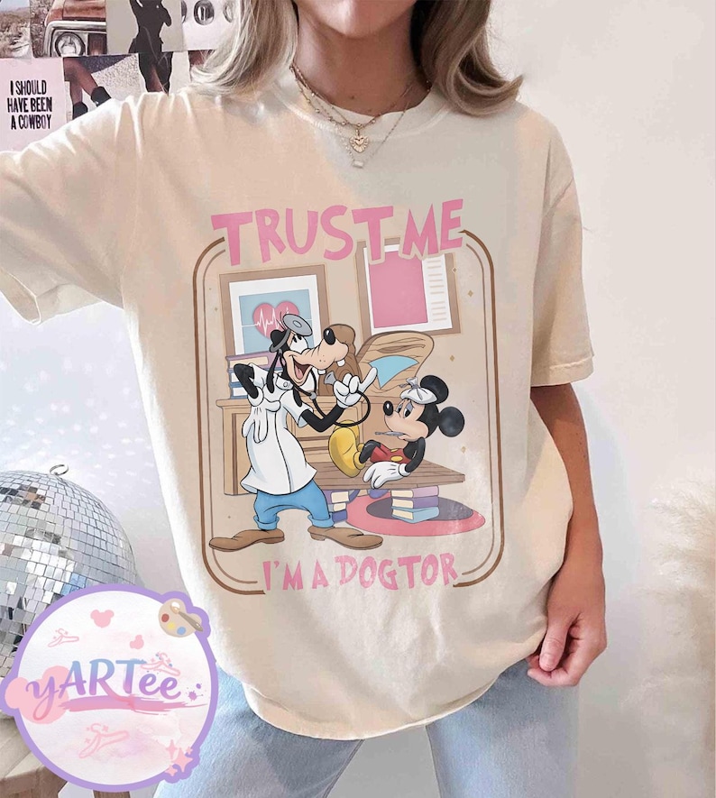 May include: Cream-coloured t-shirt with a cartoon illustration of Goofy and Mickey Mouse as doctors. The text "TRUST-ME I'M A DOGTOR" is printed in pink and brown. The shirt has a relaxed fit and is made of a soft material.