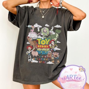 May include: Dark gray t-shirt featuring a colorful graphic of Toy Story characters, including Woody, Buzz Lightyear, and Rex. The text "TOY STORY" is prominently displayed above the characters, with "YOU'VE GOT A FRIEND IN ME" below.