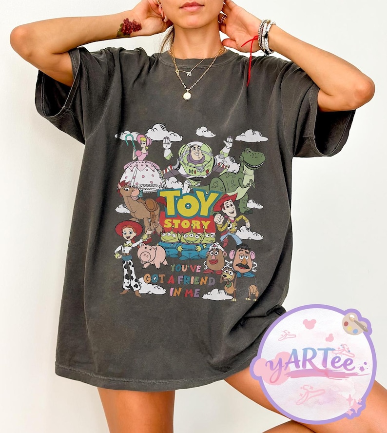 May include: Dark gray t-shirt featuring a colorful graphic of Toy Story characters, including Woody, Buzz Lightyear, and Rex. The text "TOY STORY" is prominently displayed above the characters, with "YOU'VE GOT A FRIEND IN ME" below.
