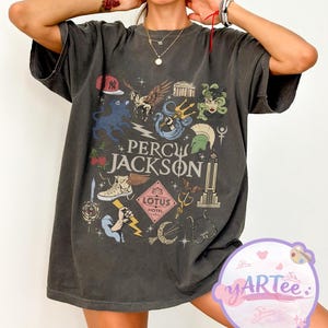 May include: Dark gray t-shirt featuring a colorful graphic design with the text "Percy Jackson" and various illustrations. The design includes a baseball cap, Pegasus, a trident, Medusa, and other themed elements. The shirt has short sleeves.
