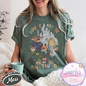 Puede incluir: Camiseta verde musgo con un gr&aacute;fico colorido de personajes de dibujos animados y el texto "WHERE ANYONE CAN BE ANYTHING". El dise&ntilde;o incluye un paisaje urbano y varias figuras de animales. La camiseta es de la colecci&oacute;n Comfort Colors.