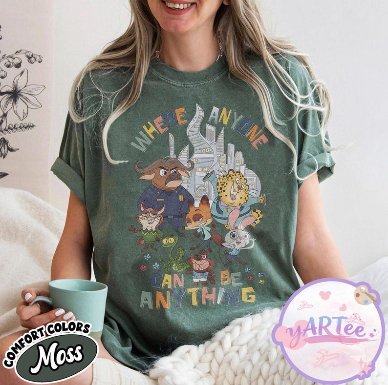 Puede incluir: Camiseta verde musgo con un gr&aacute;fico colorido de personajes de dibujos animados y el texto "WHERE ANYONE CAN BE ANYTHING". El dise&ntilde;o incluye un paisaje urbano y varias figuras de animales. La camiseta es de la colecci&oacute;n Comfort Colors.