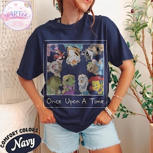 May include: Navy blue t-shirt featuring a colorful graphic of cartoon characters from various animated films. The graphic is framed with a white border and includes the text "Once Upon A Time". The shirt is labeled "Comfort Colors Navy".