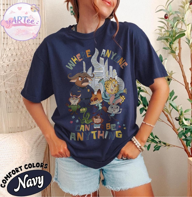 Puede incluir: Camiseta azul marino con un gr&aacute;fico colorido con personajes de dibujos animados y el texto "WHERE ANYONE CAN BE ANYTHING". La camiseta tiene cuello redondo y mangas cortas. La marca "YARtee" es visible en la esquina superior izquierda.