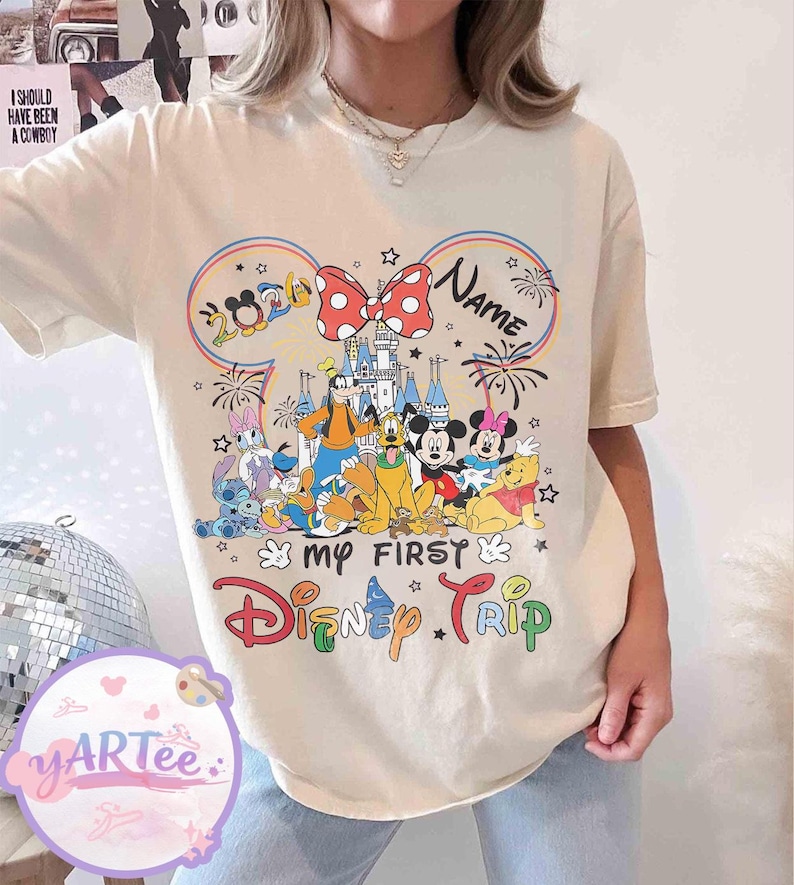 May include: Cream-colored t-shirt featuring a colorful graphic of Disney characters, including Mickey and Minnie Mouse, Goofy, and Winnie the Pooh. The design includes the text "My First Disney Trip" and "2024". A red bow with white polka dots is also featured.