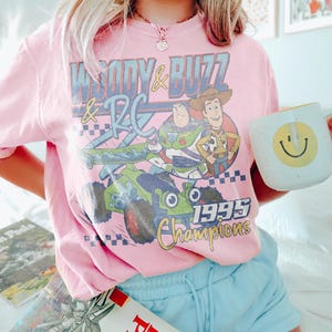 May include: Pink t-shirt featuring Woody, Buzz Lightyear, and RC from Toy Story, with the text "Woody & Buzz & RC 1995 Champions." The shirt is paired with light blue shorts and a white mug with a yellow smiley face.