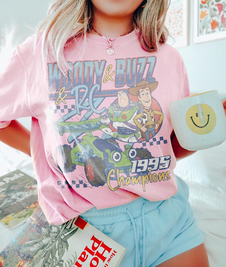 May include: Pink t-shirt featuring Woody, Buzz Lightyear, and RC from Toy Story, with the text "Woody & Buzz & RC 1995 Champions." The shirt is paired with light blue shorts and a white mug with a yellow smiley face.