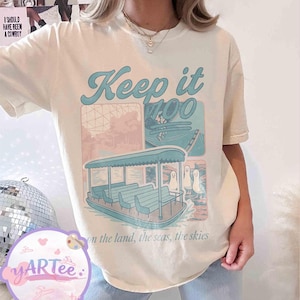 May include: Cream-colored t-shirt with a graphic featuring a boat, the text "Keep it 100", and the phrase "on the land, the seas, the skies". The design includes pastel blue and pink elements.