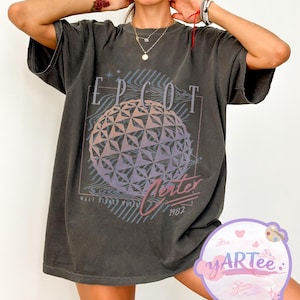 May include: Dark gray t-shirt featuring the word "EPCOT" above a geometric sphere design in shades of purple and pink. Below the sphere, the word "Center" is written in a cursive font, with "Walt Disney World" and the year "1982" below.