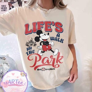 May include: Cream-coloured t-shirt with Mickey Mouse walking, and the text "LIFE'S A WALK IN THE PARK" in red and blue. The Walt Disney World logo is at the bottom. A castle graphic is also included.