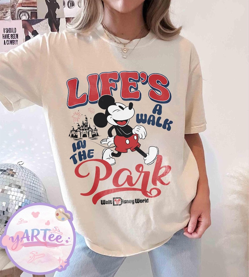 May include: Cream-coloured t-shirt with Mickey Mouse walking, and the text "LIFE'S A WALK IN THE PARK" in red and blue. The Walt Disney World logo is at the bottom. A castle graphic is also included.