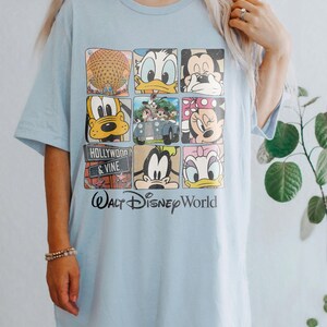 Op de afbeelding: Lichtblauw T-shirt met een grafische print van verschillende Disney-figuren, waaronder Mickey Mouse, Donald Duck, Goofy en Pluto. Het shirt heeft ook de tekst "Walt Disney World" en "Hollywood & Vine".