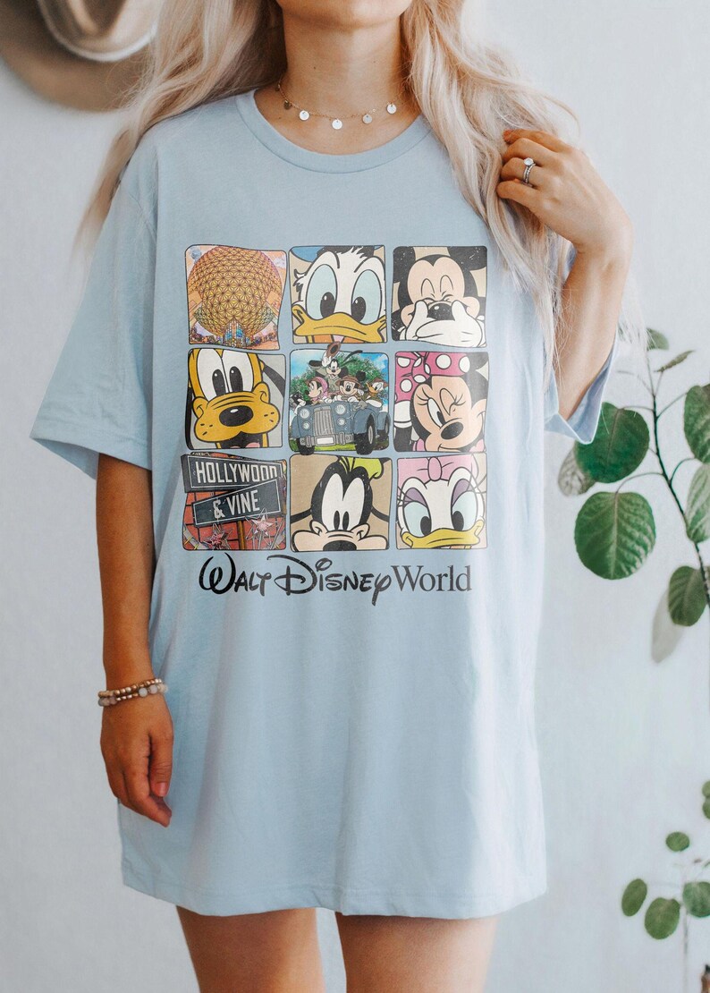 Op de afbeelding: Lichtblauw T-shirt met een grafische print van verschillende Disney-figuren, waaronder Mickey Mouse, Donald Duck, Goofy en Pluto. Het shirt heeft ook de tekst "Walt Disney World" en "Hollywood & Vine".
