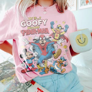 May include: A pale pink t-shirt with a graphic of Goofy as Spider-Man, alongside Mickey Mouse, Donald Duck, and other characters. The shirt displays the text "WHAT IF...? GOOFY BECAME SPIDER-MAN". A white mug with a yellow smiley face is held in the hand.