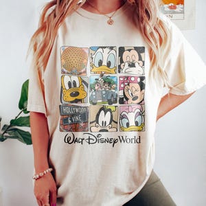 Op de afbeelding: Een beige T-shirt met een grafische print van verschillende Disney-figuren, waaronder Mickey Mouse, Minnie Mouse, Donald Duck, Goofy en Pluto. De afbeelding bevat ook een foto van de Spaceship Earth-attractie in Epcot en een bord met de tekst "Hollywood & Vine". De tekst "Walt Disney World" staat onder de afbeelding.