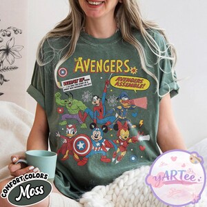 May include: A moss green t-shirt featuring a comic-style graphic of The Avengers characters, including Hulk, Spider-Man, and Mickey Mouse. The shirt has the text "The Avengers" and "Avengers Assemble!" printed on it. The shirt is made of Comfort Colors material.