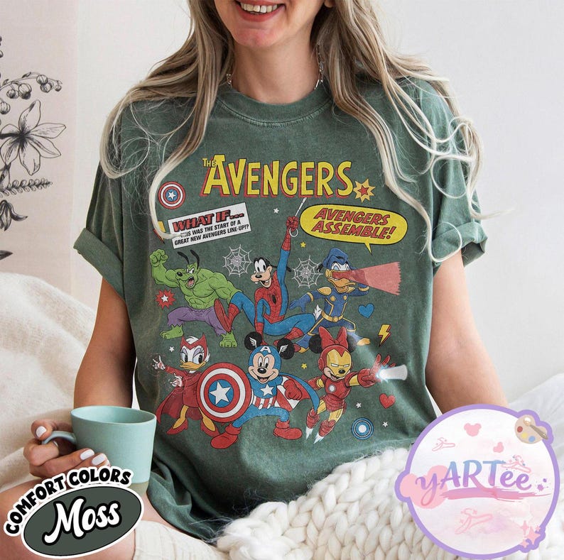 May include: A moss green t-shirt featuring a comic-style graphic of The Avengers characters, including Hulk, Spider-Man, and Mickey Mouse. The shirt has the text "The Avengers" and "Avengers Assemble!" printed on it. The shirt is made of Comfort Colors material.