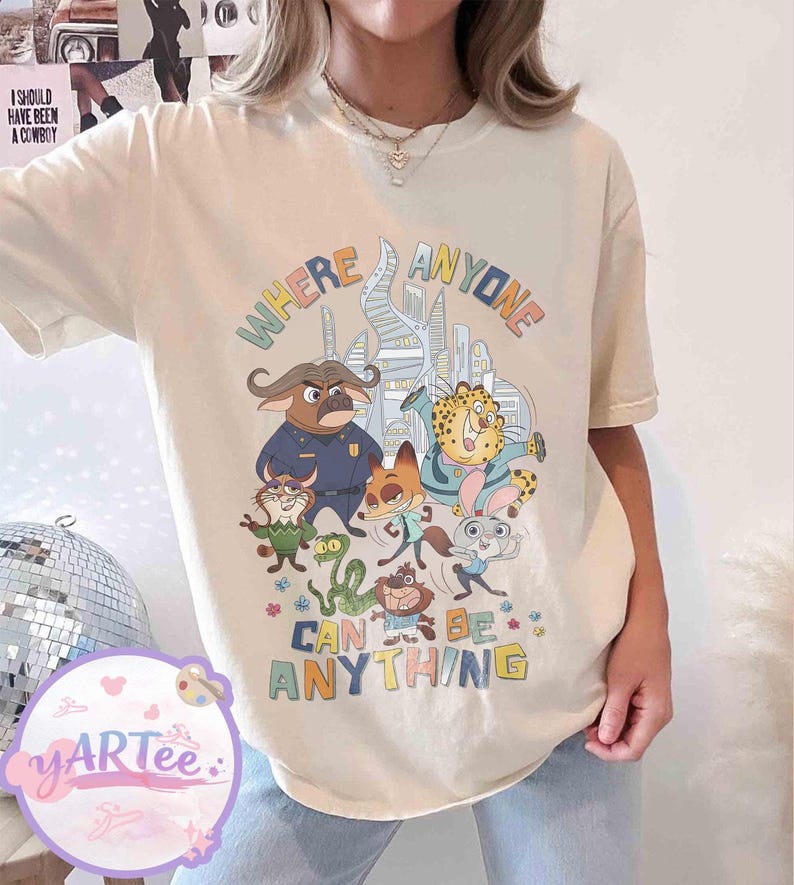 Puede incluir: Camiseta color crema con un gr&aacute;fico colorido de personajes de dibujos animados y el texto "WHERE ANYONE CAN BE ANYTHING". El dise&ntilde;o presenta personajes de animales en un entorno urbano, con un estilo l&uacute;dico y caprichoso.