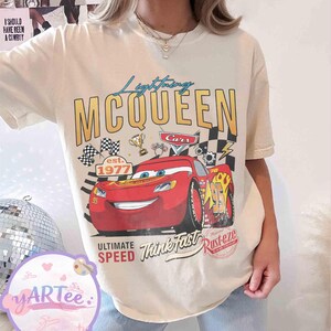 Pu&ograve; includere: T-shirt color crema con grafica di Saetta McQueen dal film Cars. Il design include il testo "Lightning McQueen", "Cars", "Ultimate Speed" e "Think Fast". La maglietta presenta anche una bandiera a scacchi e la scritta "est. 1977."