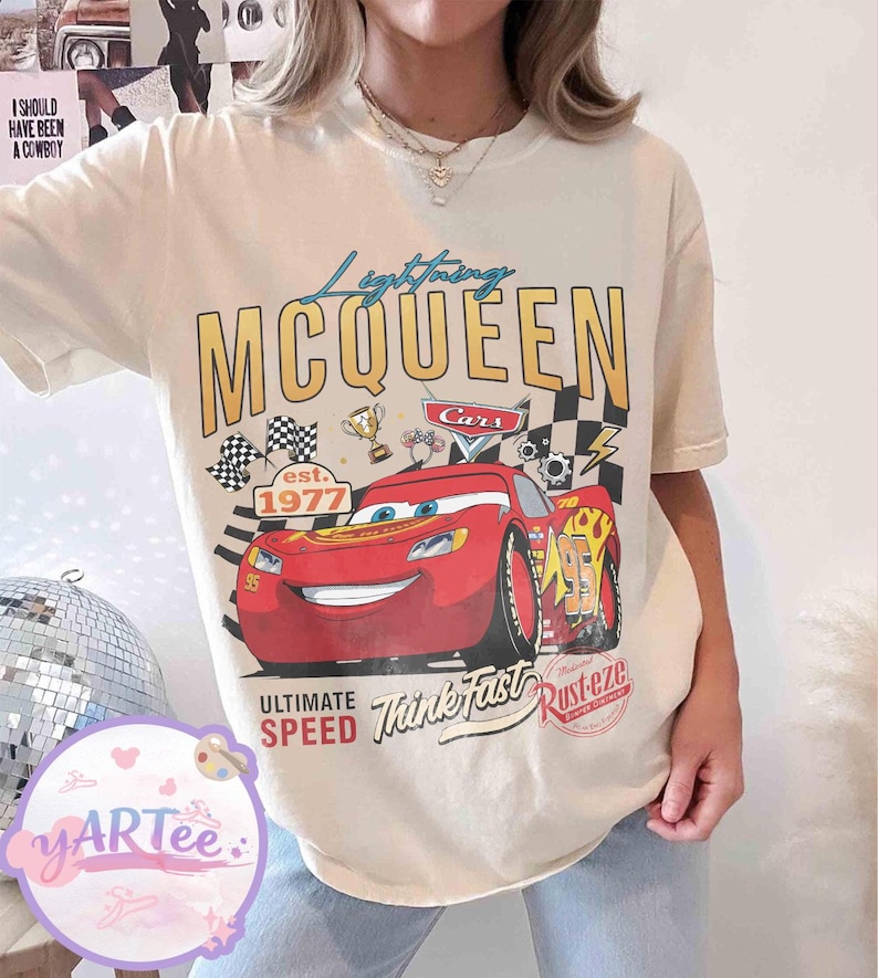 Pu&ograve; includere: T-shirt color crema con grafica di Saetta McQueen dal film Cars. Il design include il testo "Lightning McQueen", "Cars", "Ultimate Speed" e "Think Fast". La maglietta presenta anche una bandiera a scacchi e la scritta "est. 1977."