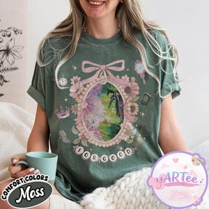 K&ouml;nnte beinhalten: Moosgr&uuml;nes T-Shirt mit einem gerahmten Grafikmotiv, das eine hexenartige Figur und den Text "FOR GOOD" zeigt. Der Rahmen ist mit Blumen, einer Krone und einem Spiegel verziert. Das Shirt tr&auml;gt auch die Aufschrift "COMFORT COLORS" und "Moss".