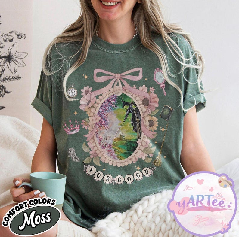 K&ouml;nnte beinhalten: Moosgr&uuml;nes T-Shirt mit einem gerahmten Grafikmotiv, das eine hexenartige Figur und den Text "FOR GOOD" zeigt. Der Rahmen ist mit Blumen, einer Krone und einem Spiegel verziert. Das Shirt tr&auml;gt auch die Aufschrift "COMFORT COLORS" und "Moss".