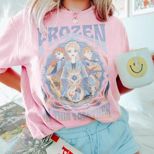 May include: Pale pink t-shirt featuring a Frozen graphic with Elsa, Anna, Kristoff, Olaf, and Sven. The text reads "FROZEN IN THIS TOGETHER". A white mug with a yellow smiley face is held in the hand.