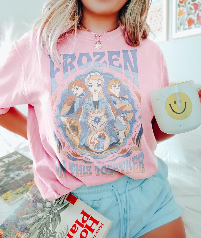 May include: Pale pink t-shirt featuring a Frozen graphic with Elsa, Anna, Kristoff, Olaf, and Sven. The text reads "FROZEN IN THIS TOGETHER". A white mug with a yellow smiley face is held in the hand.