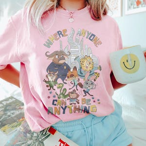 Puede incluir: Camiseta rosa claro con un gr&aacute;fico colorido que presenta personajes de animales de dibujos animados y el texto "WHERE ANYONE CAN BE ANYTHING". El dise&ntilde;o incluye un paisaje urbano y varias figuras de animales. Se sostiene una taza blanca con una carita sonriente amarilla.