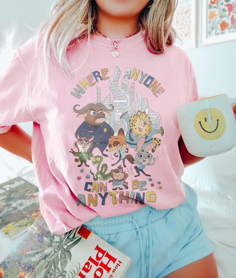 Puede incluir: Camiseta rosa claro con un gr&aacute;fico colorido que presenta personajes de animales de dibujos animados y el texto "WHERE ANYONE CAN BE ANYTHING". El dise&ntilde;o incluye un paisaje urbano y varias figuras de animales. Se sostiene una taza blanca con una carita sonriente amarilla.