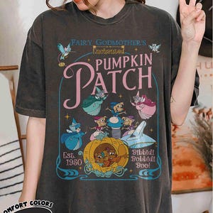 May include: Dark gray t-shirt featuring a whimsical design from "Fairy Godmother's Enchanted Pumpkin Patch." The graphic includes cartoon characters, a pumpkin carriage, and the text "Bibbidi Bobbidi Boo!" and "Est. 1950." The shirt is from Comfort Colors.